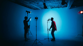 Two individuals in a creative studio setting, one adjusting equipment and the other singing into a microphone, illuminated by vibrant blue lighting, representing collaborative equipment management in media production.