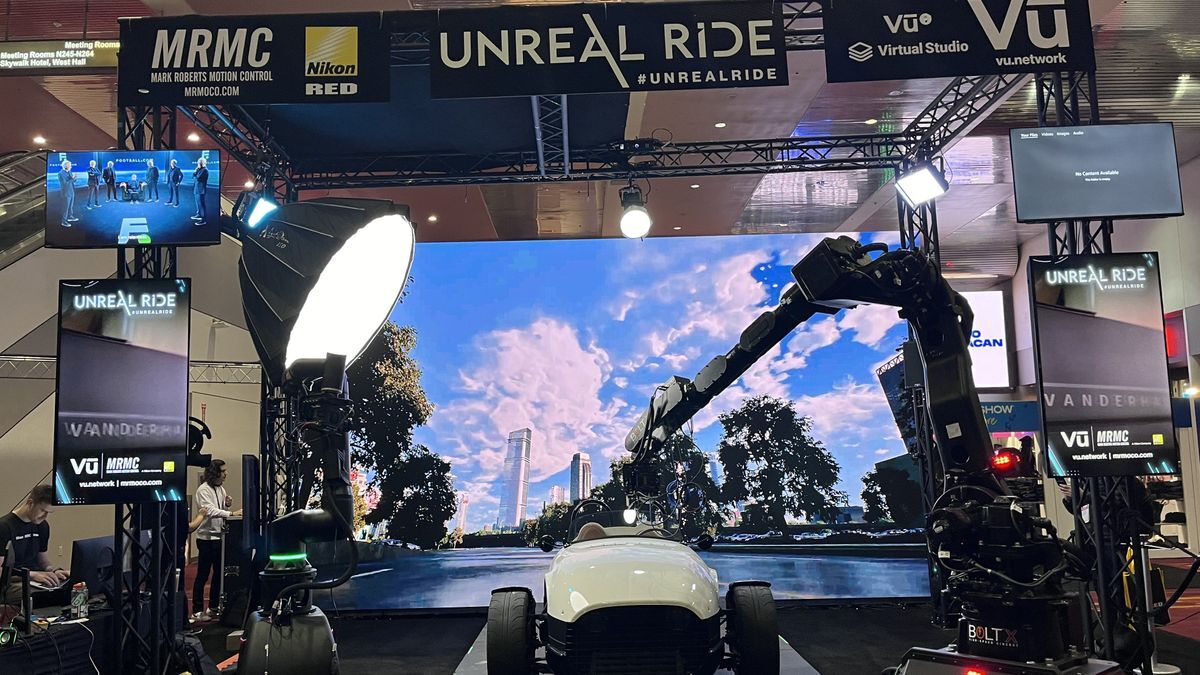 Exhibition setup featuring a white vehicle with a robotic camera arm, illuminated by studio lights, against a large digital backdrop displaying a scenic landscape, showcasing advanced motion control technology for media production.